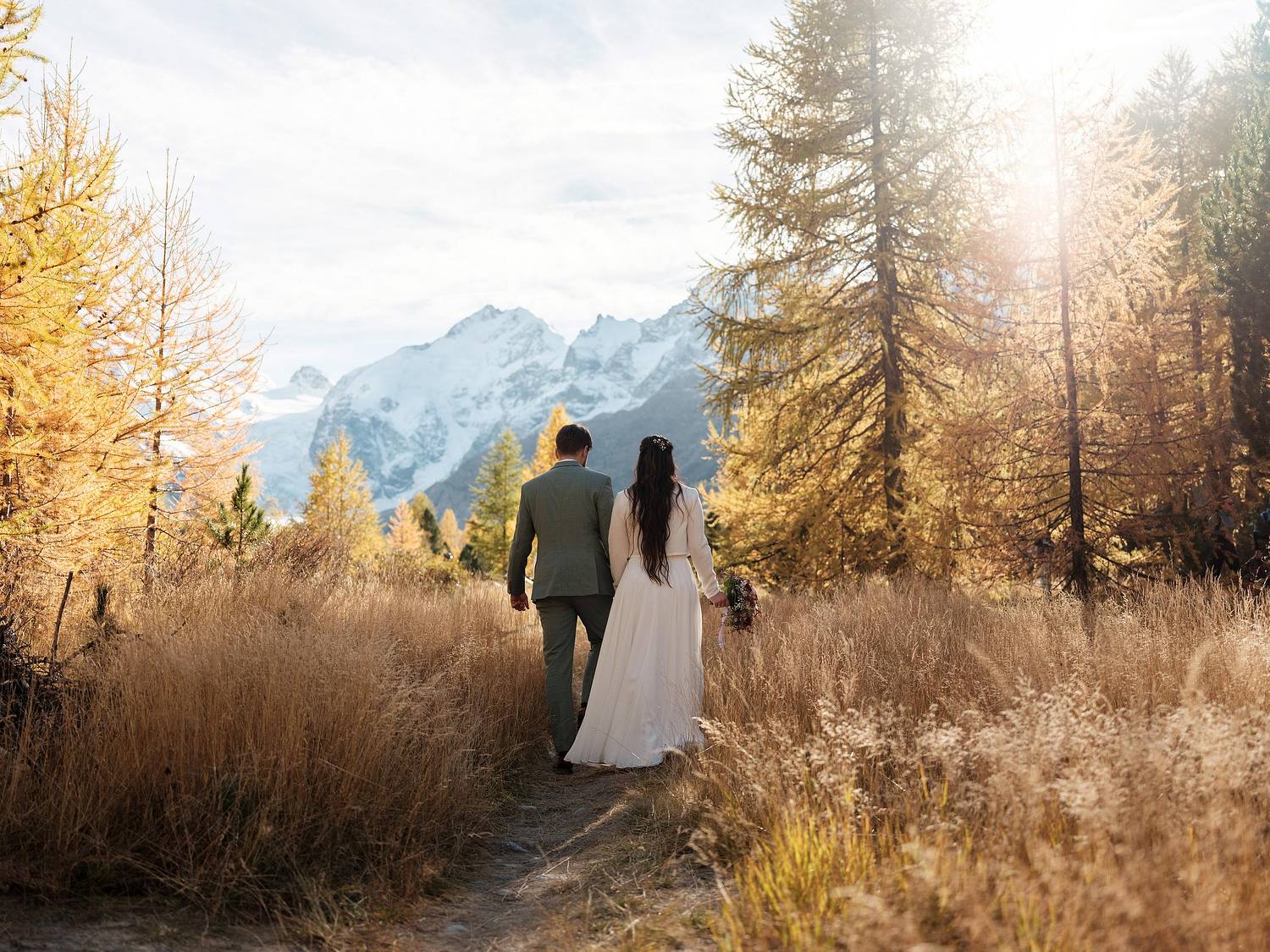 Brautpaar Simona & Christian wandert Hand in Hand durch goldene Lärchenwiesen vor dem Bernina-Gletscher, Hochzeitsfotograf Graubünden